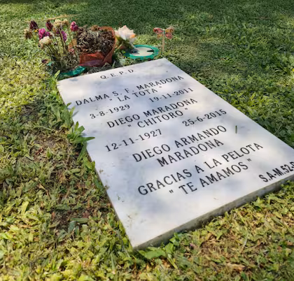 CÓMO MURIÓ MARADONA Una lápida blanca colocada en el césped muestra los nombres de Dalma Salvadora Franco, Diego Maradona y Diego Armando Maradona, con flores y una pequeña planta en la parte superior, invitando a los visitantes a reflexionar sobre CÓMO MURIÓ una de las mayores leyendas del fútbol.